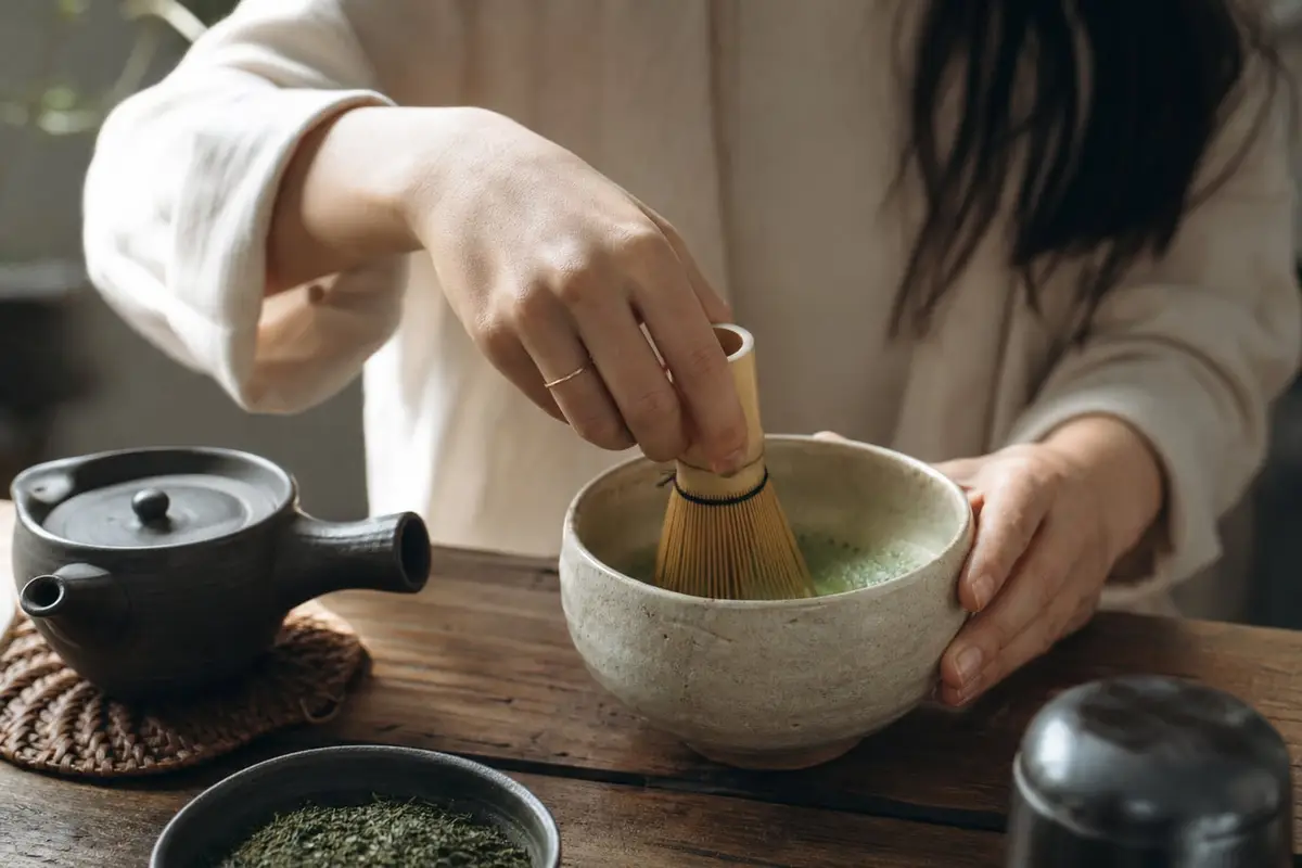 Des mains fouettent un matcha au chasen dans un bol large jusqu’à obtenir une mousse fine