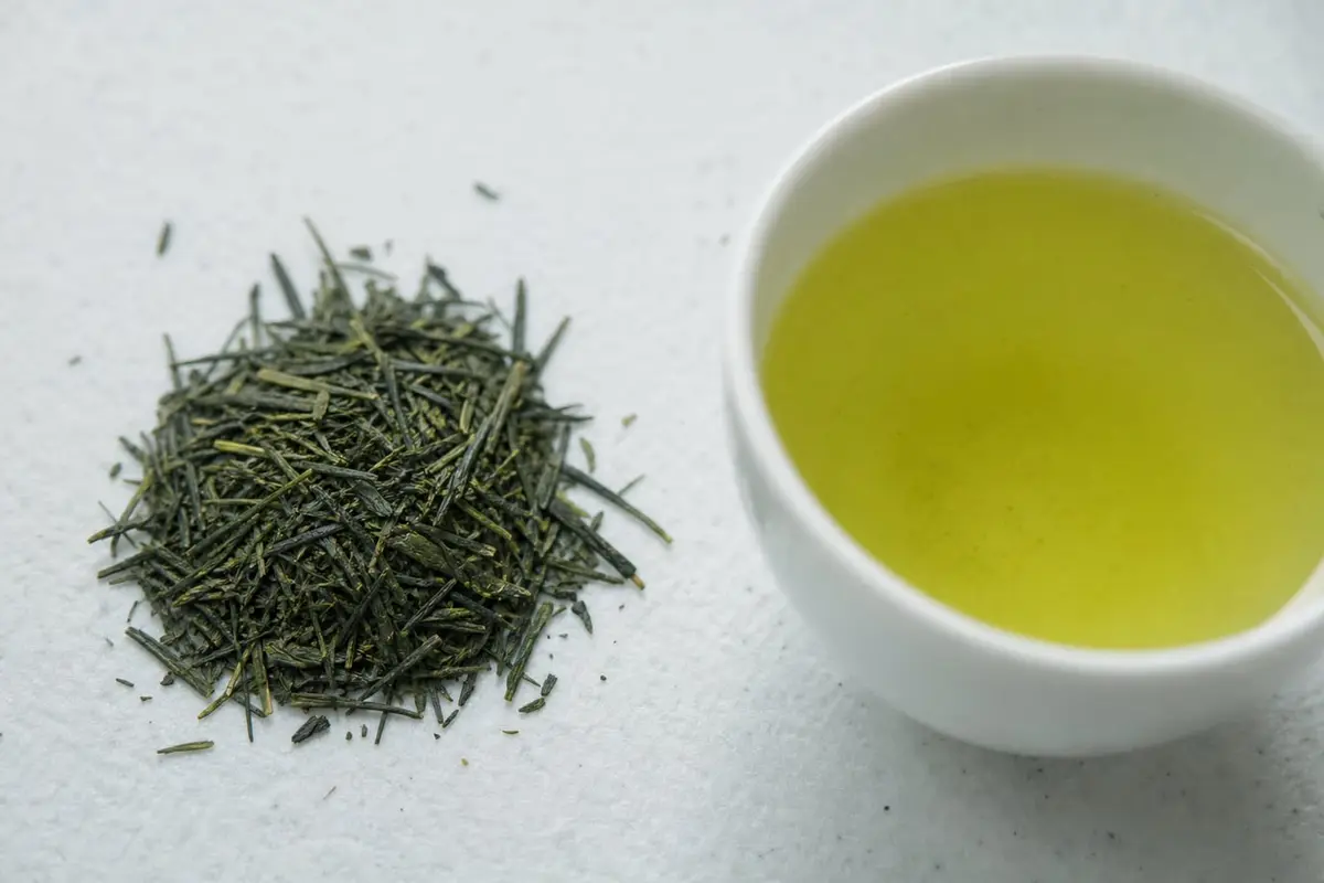 Des feuilles de sencha en aiguilles vert profond à côté d’une tasse de thé vert jaune